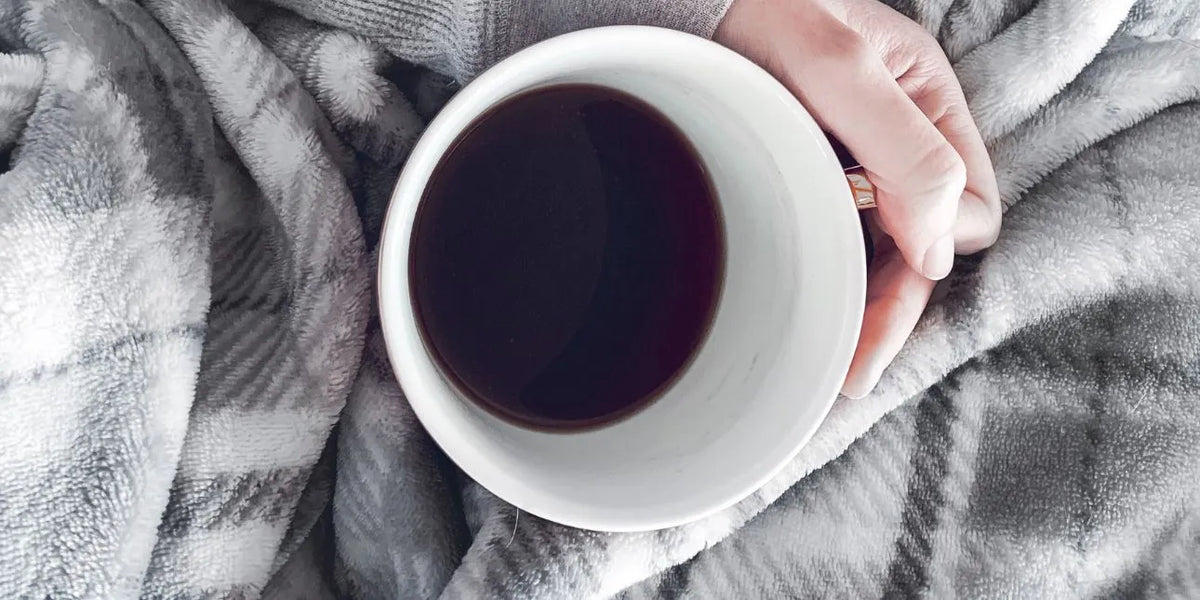 Bare biology model holding coffee mug whilst resting with blanket