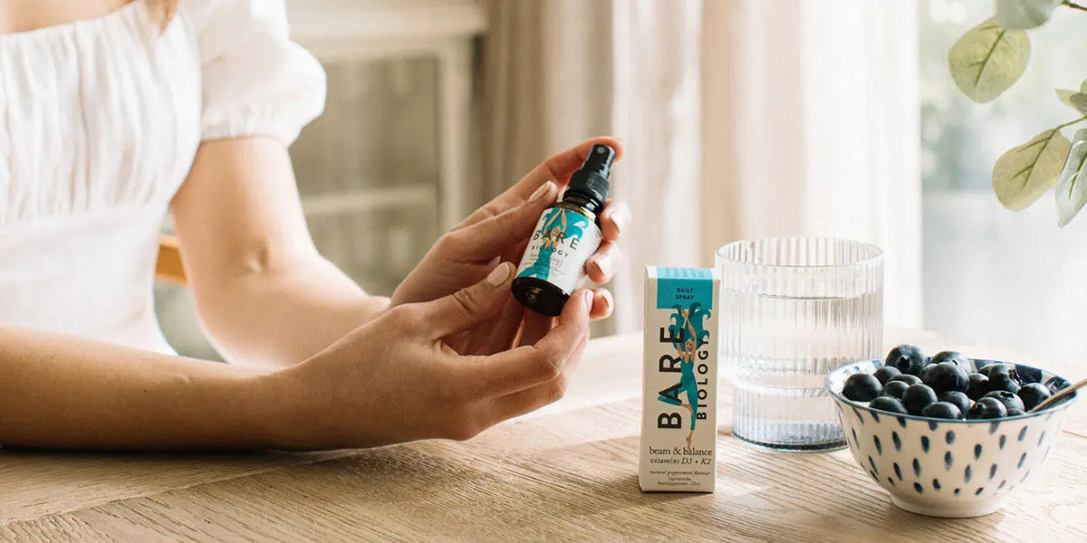 Women holding Bare Biology Beam & Balance Vitamin D3 and K2 spray at a kitchen table with a glass of water and bowl of blueberries