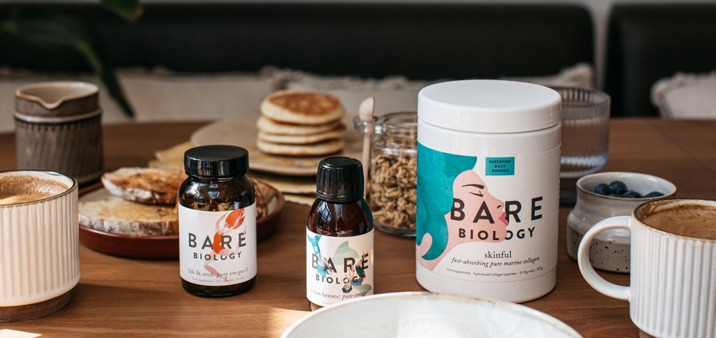 Bare Biology Skinful marine collagen, kids liquid fish oil and daily omega-3 capsules on a breakfast table amongst granola, coffee, toast and pancakes.