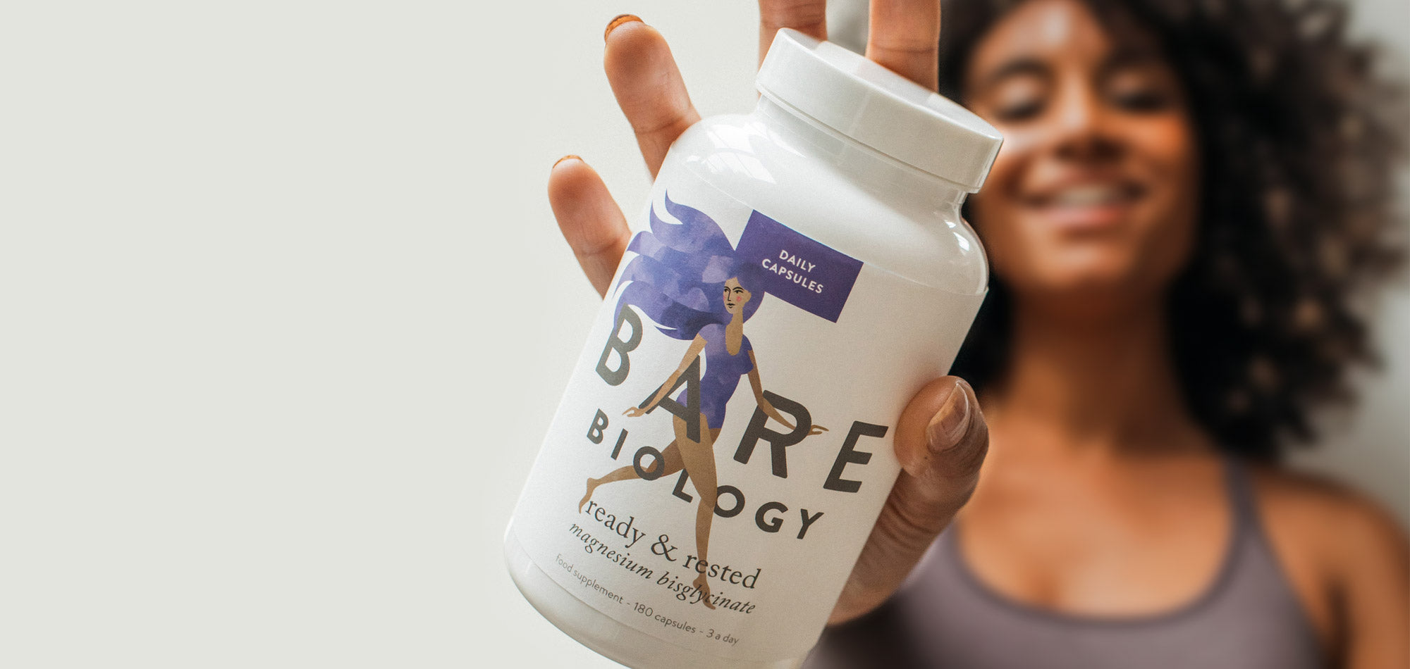 Lady blurred in the background holding an in focus jar of Bare Biology Ready & Rested magnesium capsules up to the camera