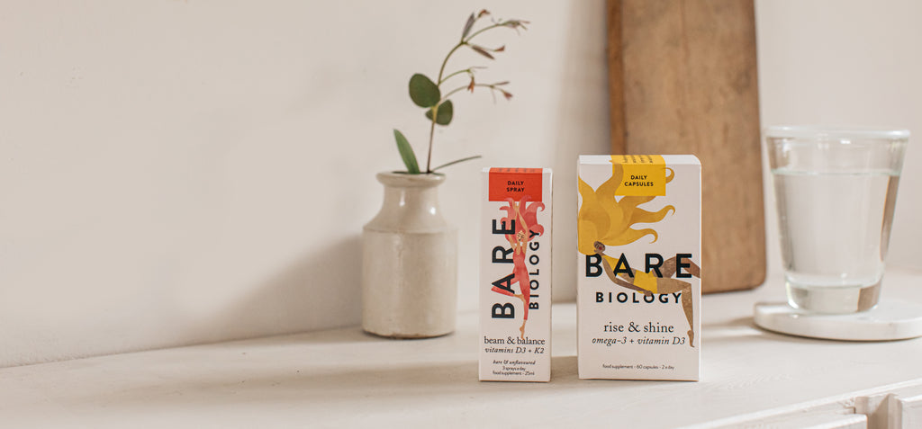 Bare Biology Rise & Shine omega-3 capsules and unflavoured vitamin D  +K2 spray on a white, wooden top next to a plant in a little vase and a glass of water.