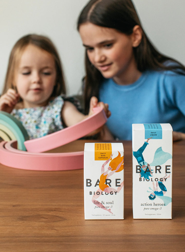 bare biology kids range life and soul mini capsules and action heroes omega 3 liquid on a table in front of a young girl supervising a toddler playing