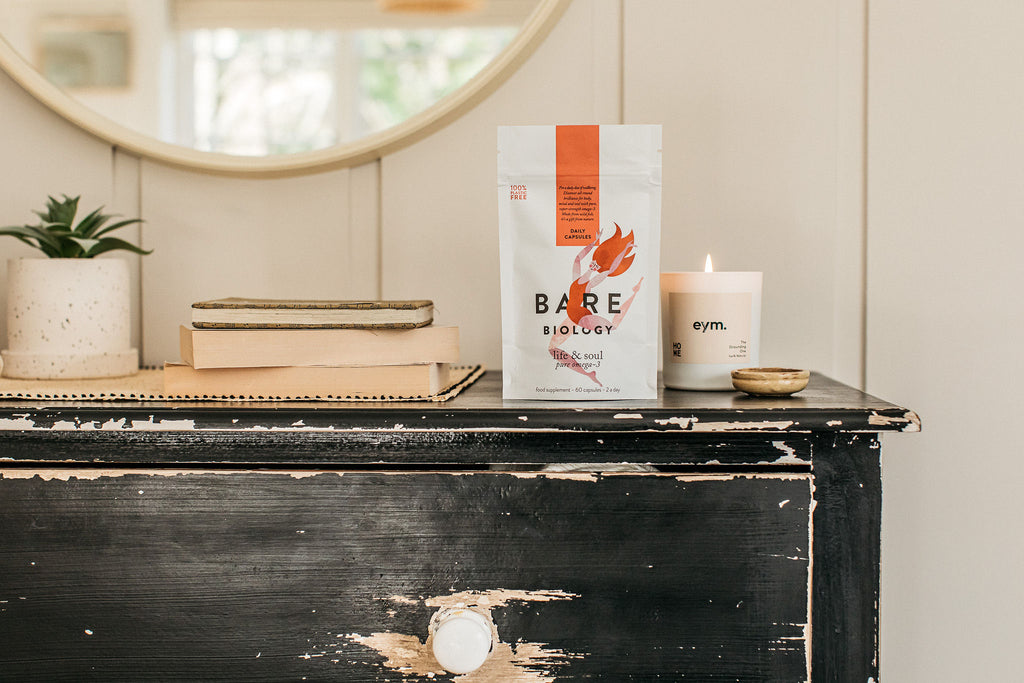 pouch of bare biology daily omega-3 capsules on a distressed black chest of drawers alongside an eym branded candle and some books.