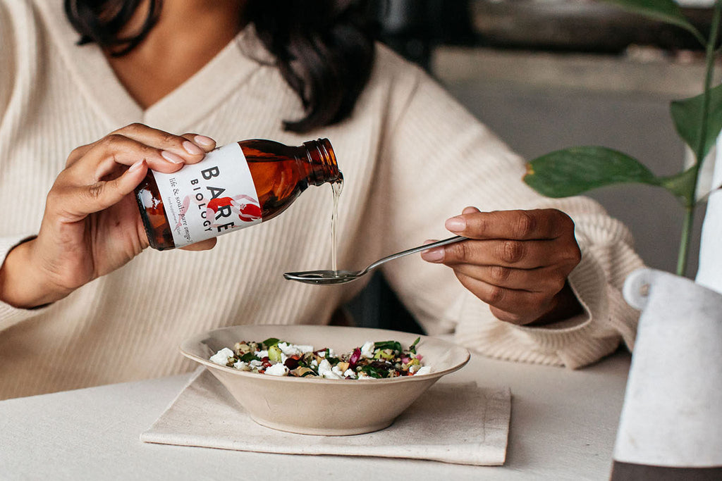 woman pouring bare biology liquid fish oil in a teaspoon over a bowl of salad.