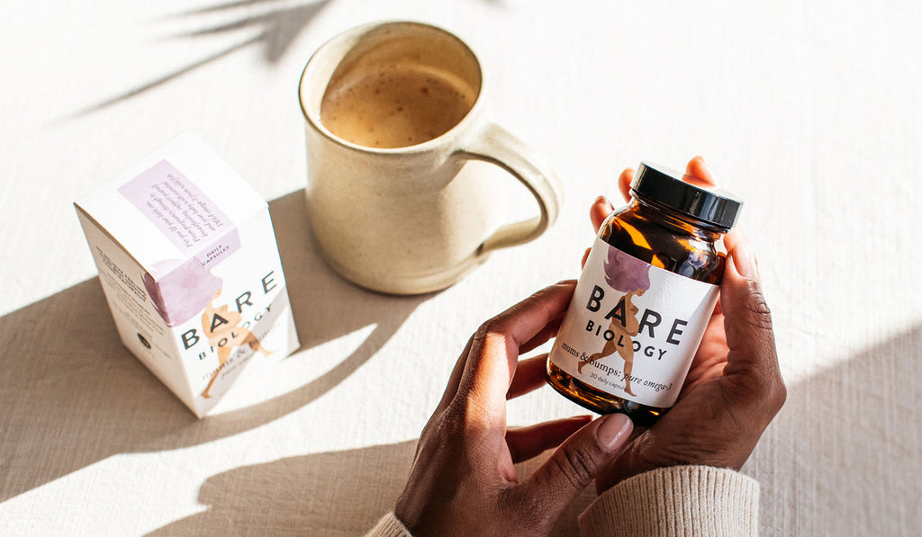 A woman's hands holding a jar of Bare Biology Mums & Bumps pregnancy omega-3 capsules, and a mug of coffee and the capsule box in the background