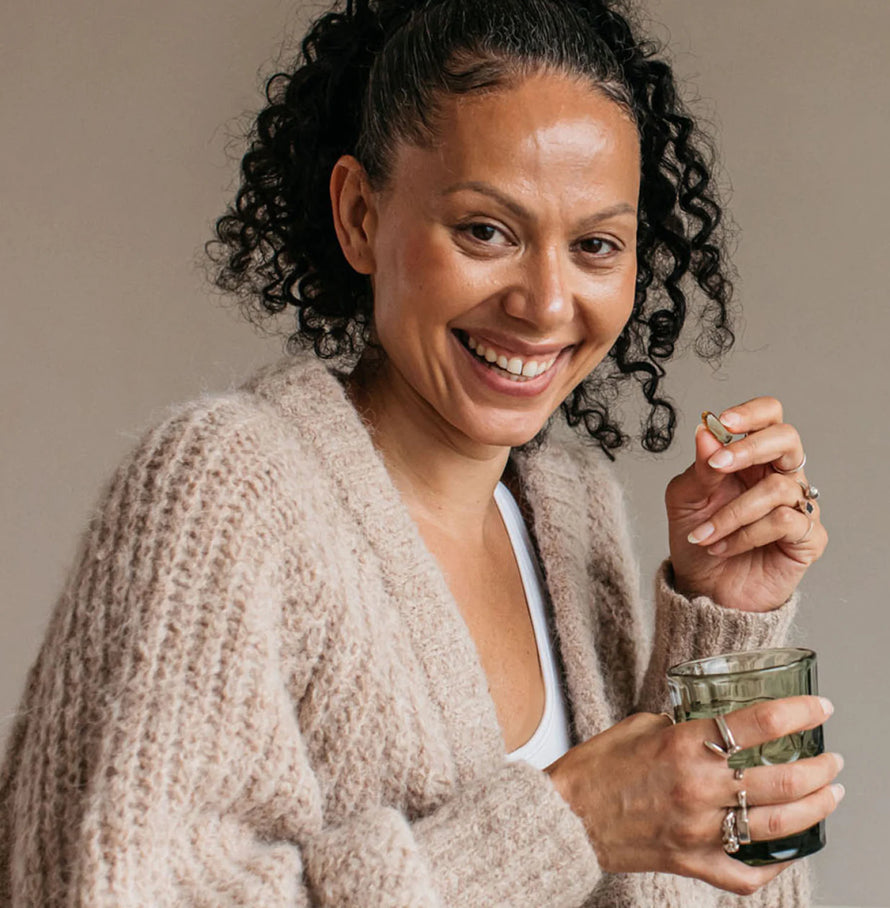 A woman in a cream jumper smiling with a glass of water and bare biology mindful omega 3 fish oil capsule