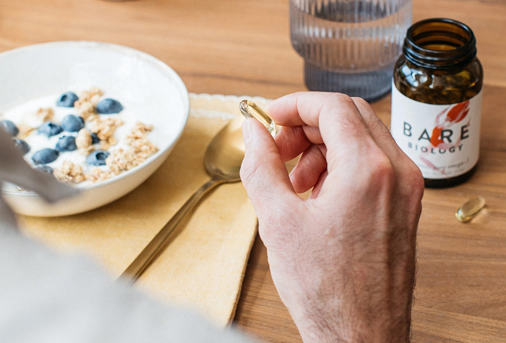 mans hand holding a bare biology omega-3 capsule with a jar of capsules and a breakfast setting in the background